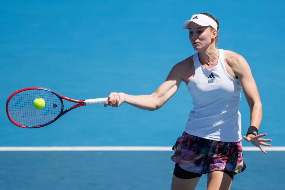 O femeie cu vizieră albă și tricou alb lovește o minge galbenă cu o rachetă de tenis pe un teren de culoare albastră sub un cer senin.