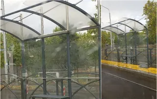 O structură de așteptare pentru transportul în comun cu acoperiș curbat și panouri de sticlă este situată lângă un peron cu bordură galbenă.