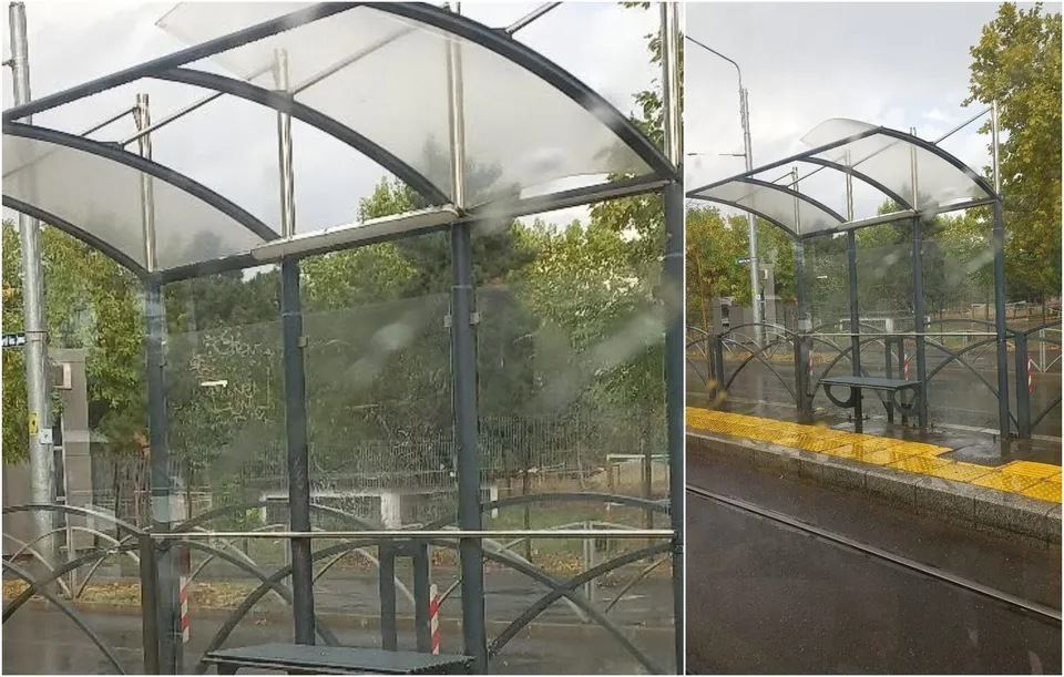 O structură de așteptare pentru transportul în comun cu acoperiș curbat și panouri de sticlă este situată lângă un peron cu bordură galbenă.