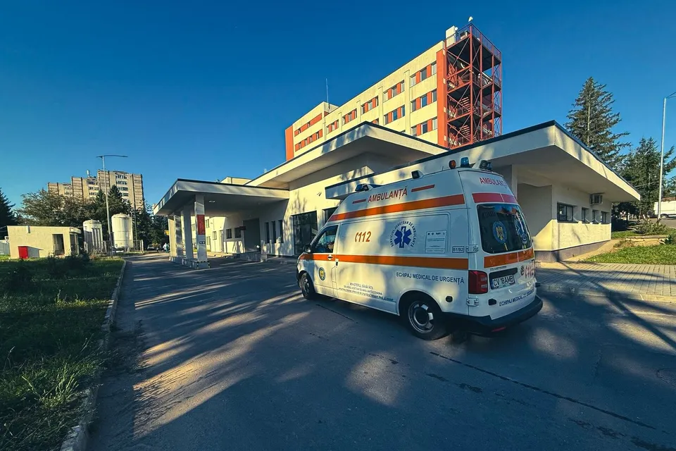 O ambulanță albă cu marcaje portocalii și un semn albastru este parcată în fața unei clădiri de spital sub un cer senin.