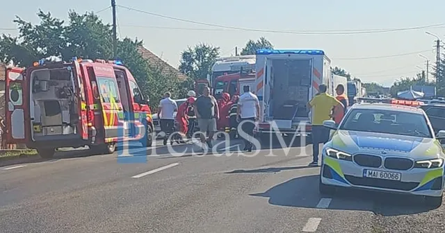 Vehicule de serviciu de urgență, inclusiv o ambulanță și un camion de pompieri, sunt parcate pe un drum lângă un grup de persoane.