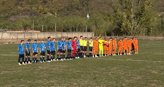 O echipă de fotbaliști stă în rând pe un teren de fotbal, purtând echipamente de culori albastre, roșii și portocalii.