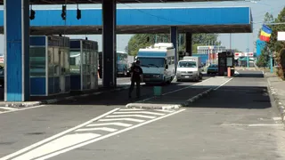 Un agent de poliție stă pe drumul asfaltat lângă două cabine de controale și un autocar alb sub o structură albastră.