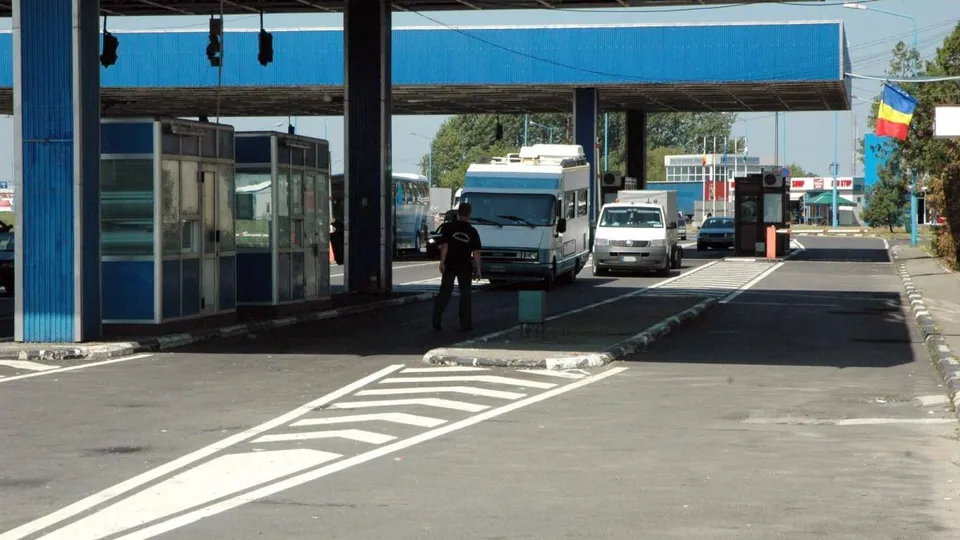 Un agent de poliție stă pe drumul asfaltat lângă două cabine de controale și un autocar alb sub o structură albastră.