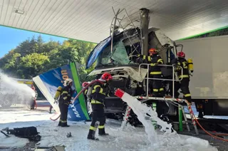 Patru pompieri în echipament de protecție folosesc furtunuri cu spumă albă pentru a stinge un incendiu la un camion aflat sub o structură de acoperiș.