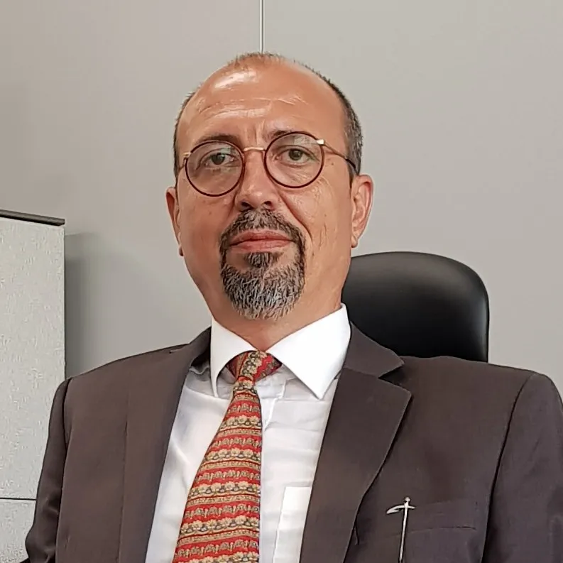 Un bărbat cu ochelari, barbă scurtă și cămașă albă cu cravată roșie stă în fața unui fundal gri.