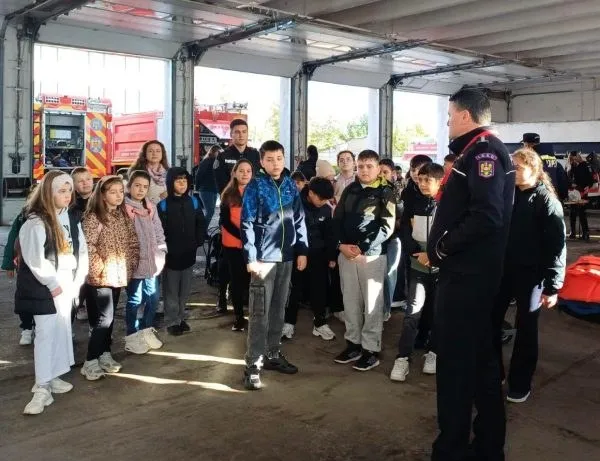 Un grup de copii stă în interiorul unui garaj deschis, ascultând un bărbat îmbrăcat în uniformă de serviciu.