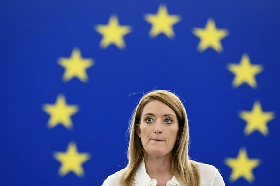 O femeie cu părul blond și bluză albă stă în fața unui fundal albastru cu stele galbene.