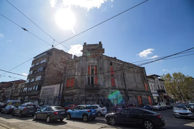 O clădire gri cu ferestre înalte și detalii arhitecturale se află lângă un bloc de apartamente, având mașini parcate în prim-plan sub un cer însorit.
