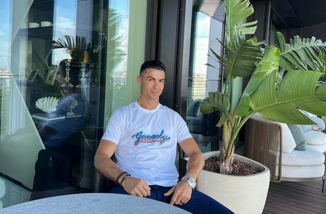 Cristiano Ronaldo stă așezat pe o terasă cu o masă rotundă în față, purtând o tricou alb cu inscripție și pantaloni albaștri, lângă o mare plantă în ghiveci.