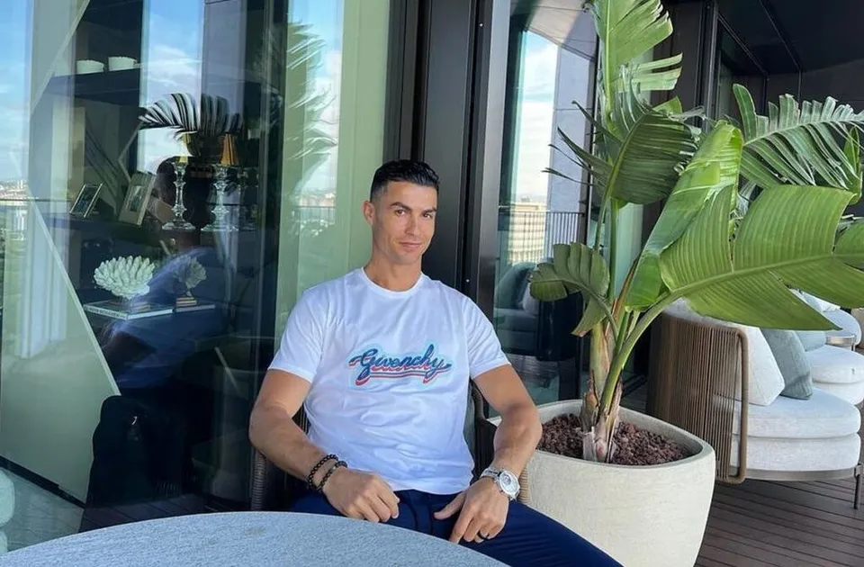 Cristiano Ronaldo stă așezat pe o terasă cu o masă rotundă în față, purtând o tricou alb cu inscripție și pantaloni albaștri, lângă o mare plantă în ghiveci.
