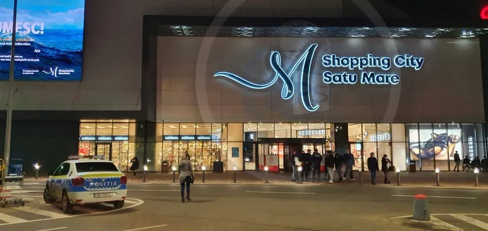 O mașină de poliție este parcată în fața unei clădiri de mall iluminată noaptea, cu oameni stând pe trotuar în apropierea intrării.