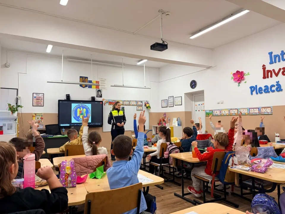 Elevii stau la bănci într-o sală de clasă și ridică mâinile în sus în timp ce o persoană stă în picioare în față.