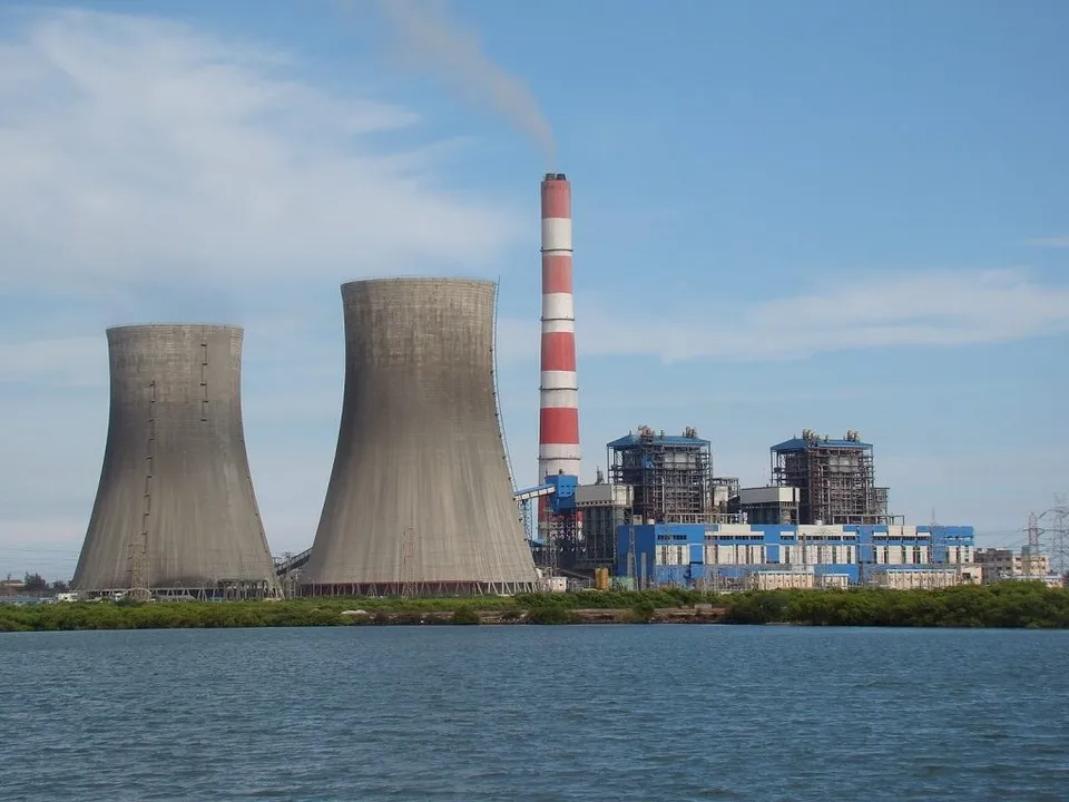 Două turnuri de răcire conice și un coș înalt cu dungi roșii și albe sunt situate lângă clădiri industriale lângă un corp de apă.