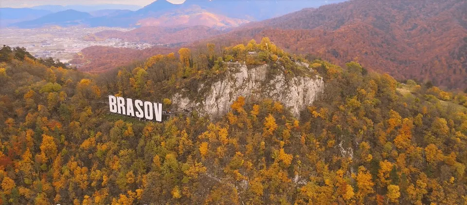 Un semn alb cu textul BRASOV este amplasat pe o versant de munte acoperit de copaci cu frunziș galben și portocaliu sub un cer înnorat.
