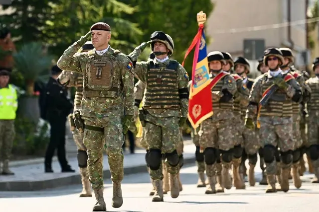 Soldați în uniformă de camuflaj și echipament tactice merg pe drum, unii salutând, în timp ce un grup poartă un steag colorat.
