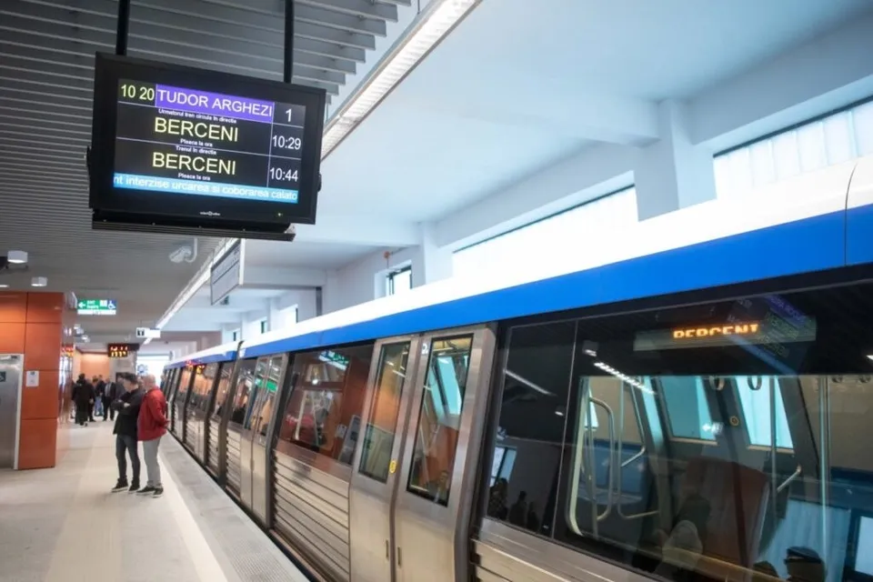 Un tren de metrou stă oprit într-o stație lângă câțiva oameni care stau pe peron sub un panou informativ suspendat.