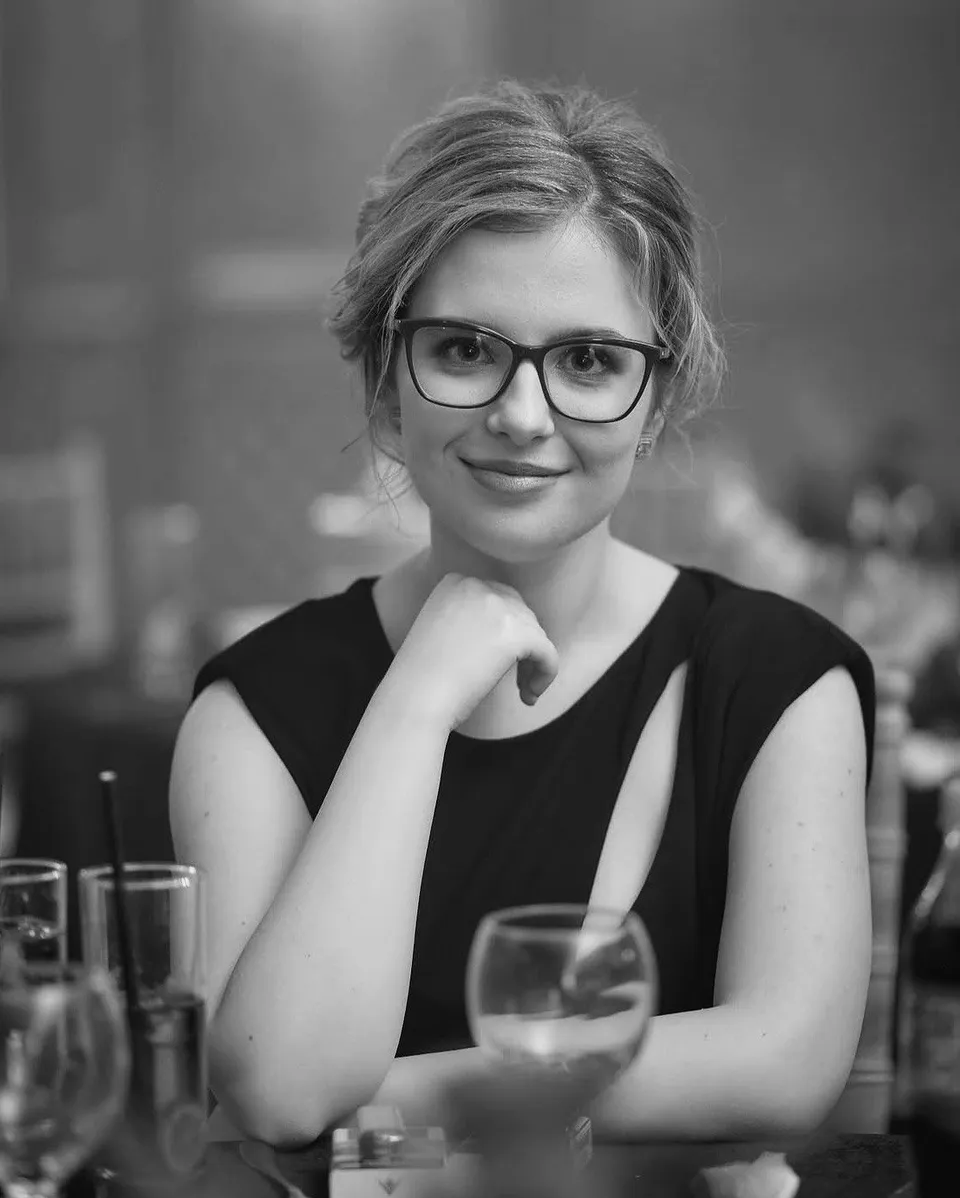 O femeie cu ochelari și părul prins într-o coafură ușoară stă cu mâna sub bărbie, purtând un tricou negru.