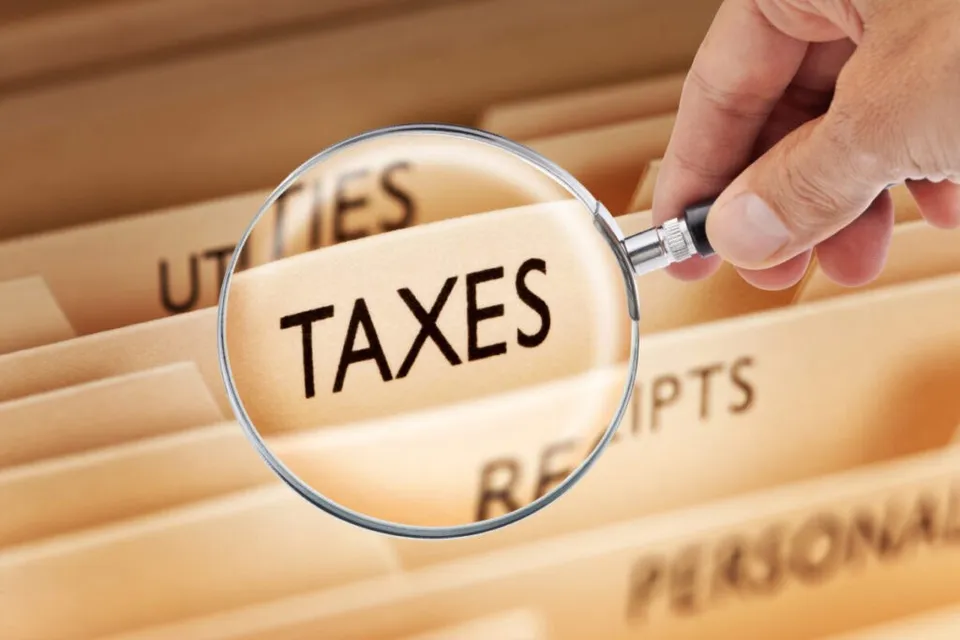 O mână ține o lupă deasupra unor dosare de arhivă pe care este scris cuvântul TAXES.