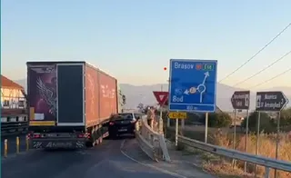 Un camion mare și un autoturism negru sunt oprite pe o porțiune de drum lângă un indicator de circulație albastru și un pod.