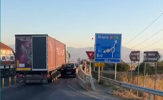 Un camion mare și un autoturism negru sunt oprite pe o porțiune de drum lângă un indicator de circulație albastru și un pod.