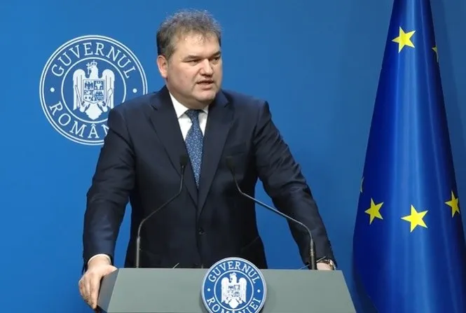 Un bărbat în costum vorbește la un pupitru, având în spate un perete albastru cu sigla Guvernului României și un steag al Uniunii Europene.