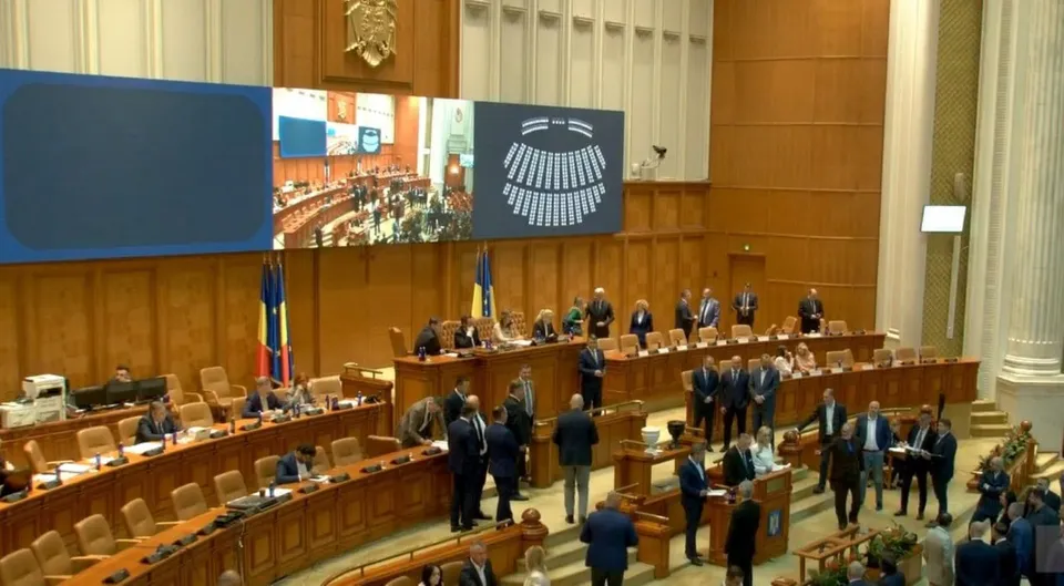 O sală de conferințe mare cu mese de lemn dispuse în rânduri, unde mai mulți oameni stau așezați sau stau în picioare.