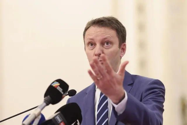 Un bărbat cu sacou albastru și cravată albastră cu dungi albe stă în fața mai multor microfoane cu mâna stângă ridicată.