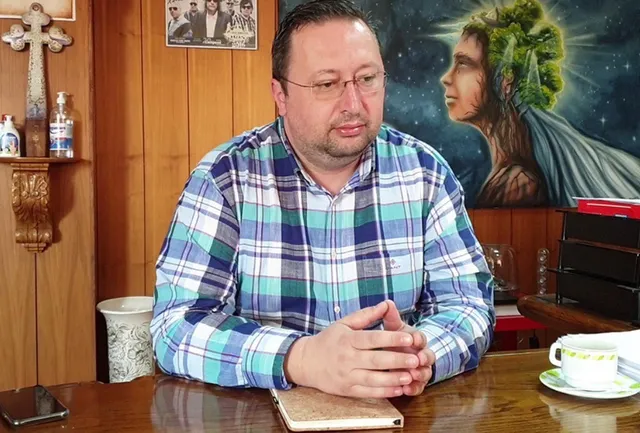 Un bărbat cu ochelari și cămașă în croituri albastre și verzi stă la o masă de lemn cu mâinile împreunate.