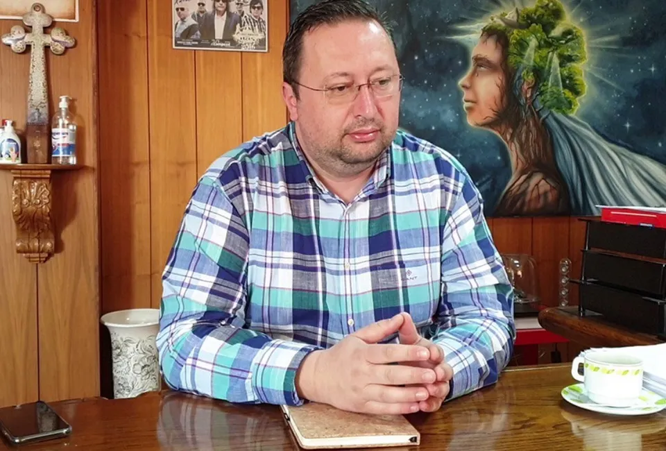 Un bărbat cu ochelari și cămașă în croituri albastre și verzi stă la o masă de lemn cu mâinile împreunate.