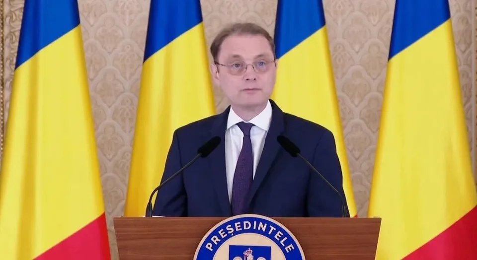 Un bărbat cu ochelari, purtând costum închis și cravată, stă în spatele unui podium în fața a patru steaguri tricolore.