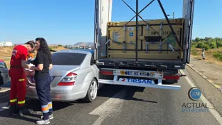 Un autoturism argintiu este blocat sub partea din spate a unui autotren care transportă un container galben pe un drum asfaltat.