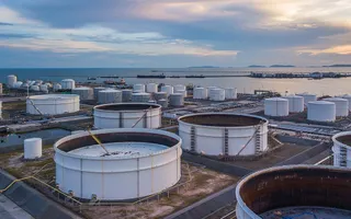 Mai multe rezervoare cilindrice albe sunt dispuse pe un teren industrial lângă o suprafață de apă sub un cer în timpul amurgului.