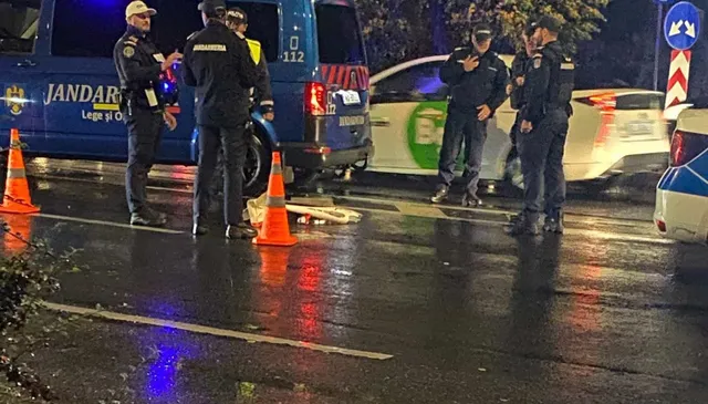 Mai mulți polițiști în uniformă stau pe un drum umed lângă un vehicul alb și o mașină de serviciu albastră cu marcaje.