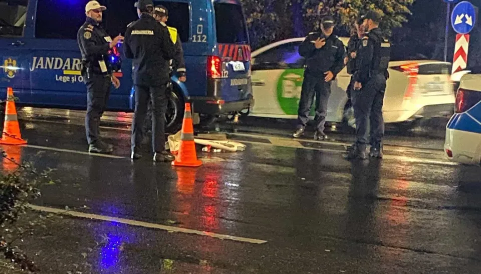 Mai mulți polițiști în uniformă stau pe un drum umed lângă un vehicul alb și o mașină de serviciu albastră cu marcaje.