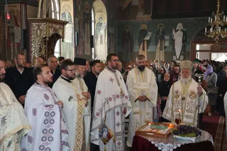 Câțiva bărbați în haine liturgice albe și decorate stau în fața unui altar cu o icoană și struguri într-o biserică cu picturi murale.