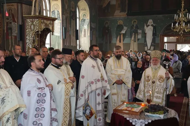 Câțiva bărbați în haine liturgice albe și decorate stau în fața unui altar cu o icoană și struguri într-o biserică cu picturi murale.
