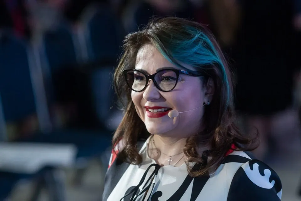 O femeie cu ochelari și păr brunet cu reflexii albastre zâmbește purtând o bluză albă cu modele negre și roșii.