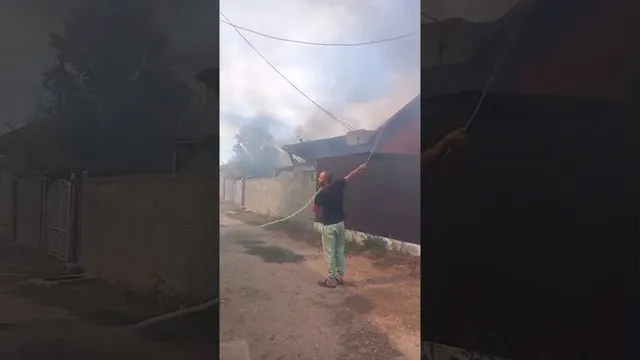 Un bărbat cu pantaloni deschiși și tricou închis de culoare ține un cablu întins deasupra unei clădiri sub un cer înnorat.