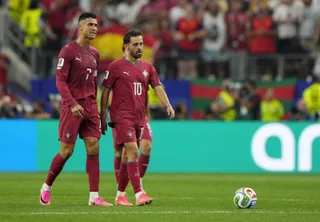 Doi jucători de fotbal îmbrăcați în echipamente de culoare roșie merg pe un teren de fotbal verde lângă un echipament de joc.