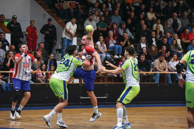 Câțiva jucători de handbal de pe un teren de culoare albastră participă la un meci în fața unui public numeros.