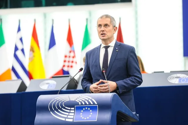 Un bărbat într-un costum albastru și cravată stă în spatele unui podium albastru cu un simbol, în fața unor steaguri colorate.