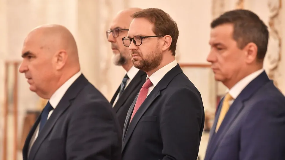 Patru bărbați în costume și cravate stau în rând, privind spre stânga într-un decor interior cu coloane și panouri decorative în fundal.