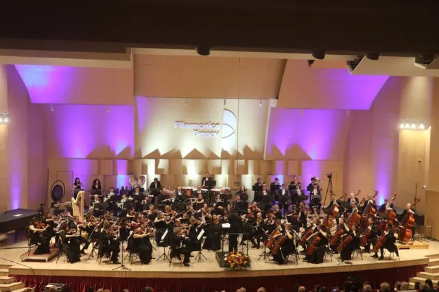 O orchestră simfonică cântă pe o scenă iluminată cu lumini mov, având instrumente precum violoncel, harpă și pian vizibile în fața publicului.