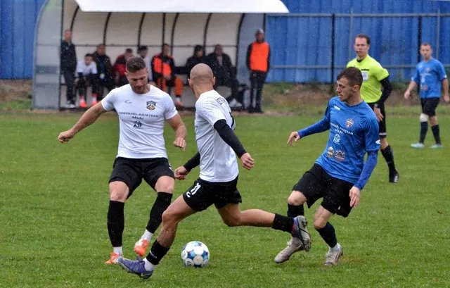 Cinci jucători de fotbal sunt pe un teren de gazon verde, unii purtând echipamente de culori albă, albastră sau galbenă.