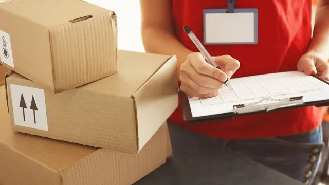 O persoană în tricou roșu scrie pe o foaie de hârtie de pe un clipboard, lângă un teanc de cutii de carton maronii.