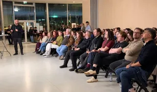 Un bărbat stă în picioare în fața unui grup de persoane așezate pe scaune într-o încăpere cu pereți deschiși spre exterior.