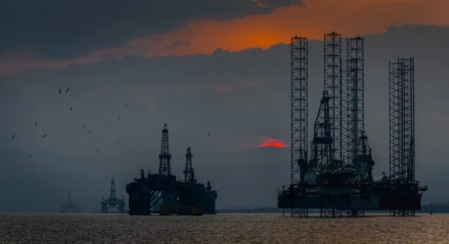 Siluete ale unor platforme de foraj petrolier situate pe apă sub un cer închis cu nuanțe portocalii la orizont.