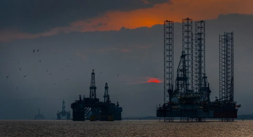 Siluete ale unor platforme de foraj petrolier situate pe apă sub un cer închis cu nuanțe portocalii la orizont.