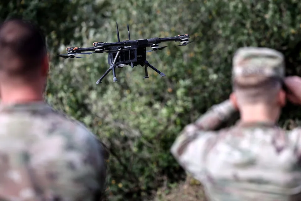 O dronă neagră zboară în aer, fiind observată de doi bărbați care poartă haine cu model camuflaj, poziționați în prim-plan.